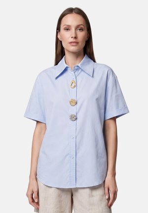 Femme portant une chemise bleu clair à manches courtes avec trois grands boutons décoratifs à l’avant, associée à un pantalon beige clair.