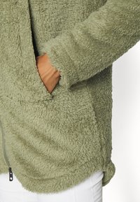 Grüne, flauschige Fleecejacke mit weicher Textur, ausgestattet mit einem Reißverschluss und Seitentaschen. Abgerundeter Saum und sichtbare Nähte.
