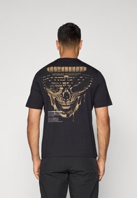 Sort t-shirt med et stort guldtryk af et kranium og tekst på ryggen. Korte ærmer og regular fit, lavet af blødt stof.