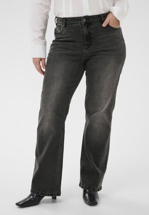 Frau trägt ausgewaschene schwarze Jeans mit geradem Bein, weißes Langarmshirt und schwarze Stiefeletten mit quadratischer Spitze und Absatz, Hand in der Tasche.