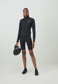 Black cycling jacket with zip, fitted design; matching black cycling shorts; holding a black helmet; sleek, lightweight material.