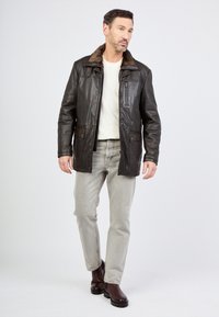 Man die loopt in een donkerbruine leren jas met een bontkraag, een wit sweater, lichtgrijze jeans en donkerbruine laarzen op een witte achtergrond.