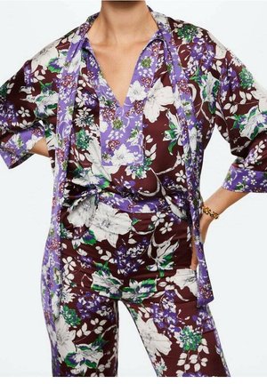 Femme porte une tenue fleurie assortie avec des fleurs violettes, blanches, vertes et marron, comprenant un haut à col en V, un pantalon et une écharpe.