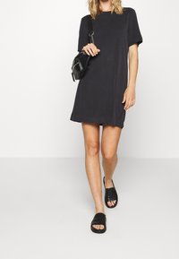 Svart t-shirtklänning med korta ärmar och avslappnad passform, tillsammans med svarta skidsandaler. Inkluderar en liten svart axelväska.