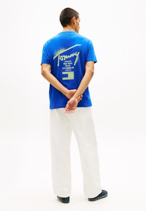 Uomo in piedi con le mani dietro la schiena, indossa una maglietta blu Tommy Jeans con nomi di città e pantaloni bianchi su uno sfondo bianco uniforme.