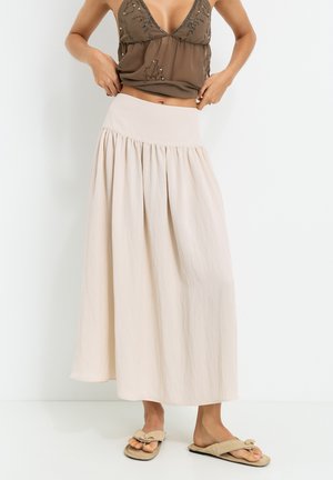 Long, flowy skirt in soft beige fabric with a wide elastic waistband and subtle pleats. Paired with a brown top and beige sandals.