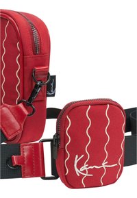 Červená látková taška přes rameno s vlnkovým bílým vzorem, vybavená zipem, nastavitelným popruhem a malou přední kapsou s logem.