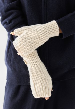 Cremefarbene, gestrickte fingerlose Handschuhe, die über dunklen, langen Ärmeln getragen werden und strukturierte Rippen- und Perlmuster zeigen.