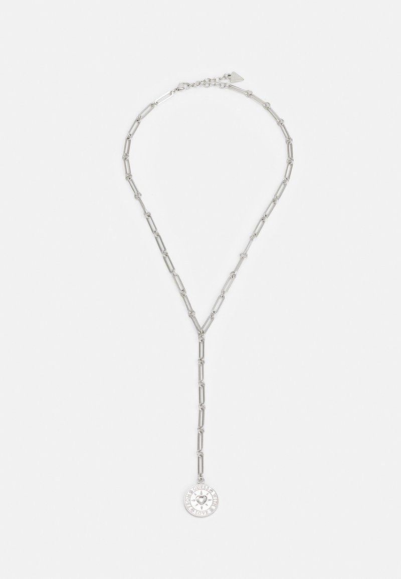 Collier chaîne en argent avec des maillons allongés et un pendentif rond gravé de "GUESS" et de symboles décoratifs, posé à plat sur un fond blanc.