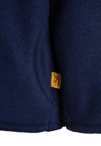 Chaqueta de forro polar azul marino con un cuello y puños acanalados. Presenta una etiqueta amarilla con texto rojo cerca de la costura. Textura suave y forma redondeada.