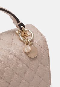 Malá béžová kabelka s prošívaným vzorem, zlatými detaily a horním uchem. Obsahuje přívěsky s logem připevněné k zipu pro ozdobu.