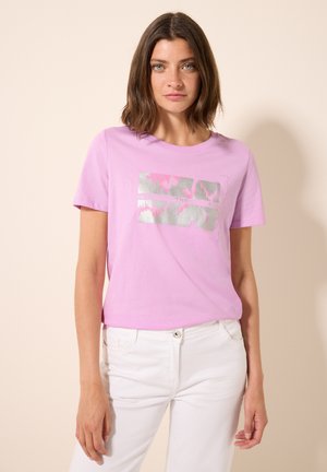 Camiseta de algodón rosa de mangas cortas, con un diseño de estampado metálico plateado. Combinada con jeans blancos, mostrando un look casual.