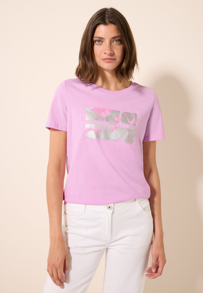 T-shirt en coton rose à manches courtes, avec un motif imprimé métallique argenté. Associé à un jean blanc, affichant un look décontracté.