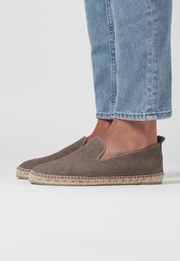 Chaussures brunes en toile tissée à enfiler avec un dessus texturé, des panneaux latéraux élastiques et des semelles en jute de style espadrille couleur beige. Portées avec un jean bleu clair.