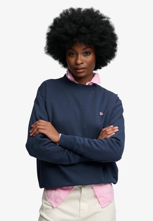 Kvinde med naturlig afro-frisure iført en mørkeblå sweatshirt over en lyserød skjorte, med armene krydset, kigger selvsikkert på kameraet.