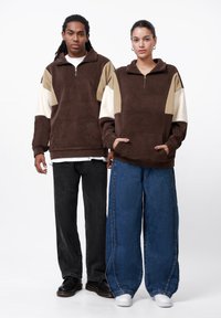 Hnedý fleece pulóver s béžovými a krémovými akcentmi, bočnými vreckami a štvrť zipsom. Osoby majú na sebe čierne a modré široké nohavice.