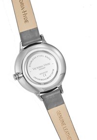 Baksidan av klockan är i silverfärgad rostfritt stål och har graverade detaljer. Remmen är i grå läder med en beige undersida som är märkt "Genuine Leather." Klockan har en rund form.