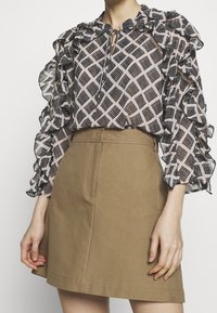 Schwarz-weiß gemusterte Bluse mit Rüsche an den Ärmeln, kombiniert mit einem hellbraunen Rock, der überfronttaschen und Steppdetails verfügt.