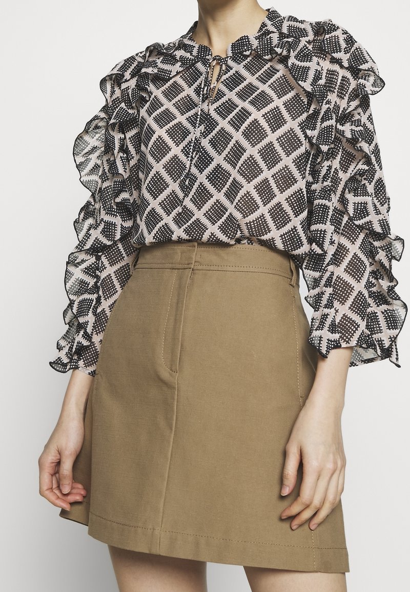 Schwarz-weiß gemusterte Bluse mit Rüsche an den Ärmeln, kombiniert mit einem hellbraunen Rock, der überfronttaschen und Steppdetails verfügt.