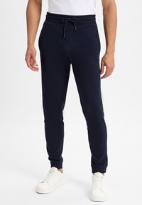 Marineblaue Jogginghosen aus weichem Stoff, mit einem elastischen Bund und Kordelzug sowie eng anliegenden Bündchen. Kombiniert mit weißen Turnschuhen.