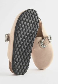 Sabots en suède beige avec un détail de boucle métallique et une semelle en caoutchouc noire à motif hexagonal pour une traction et une durabilité.