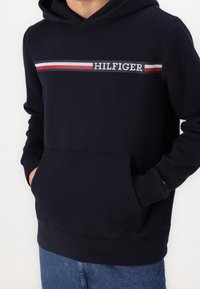 Felpa con cappuccio blu navy con tasca frontale, caratterizzata da una striscia orizzontale bianca, rossa e grigia sul petto con il testo "HILFIGER".