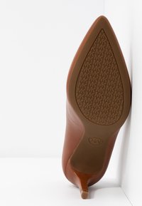 Chaussure à talon haut marron à bout pointu montrant la semelle estampillée d'un motif répété "MK" avec un petit embout de talon.