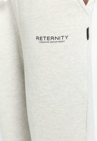 Helle graue Jogginghosen aus weichem Stoff, mit einer Seitentasche und "RETERNITY CREATIVE DEPARTMENT" in schwarzer Schrift auf dem Oberschenkel.