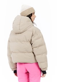 Beige pufferjack met een cropped ontwerp, grote capuchon en elastische boorden, gecombineerd met roze broek. De stof lijkt glad en geïsoleerd.