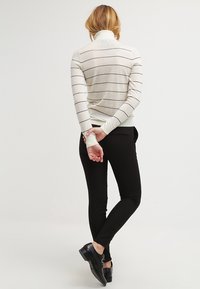 Krémový svetr s černými pruhy, rolák, žebrované manžety; kombinovaný s těsnými černými kalhotami a černými botami. Pohled zezadu.