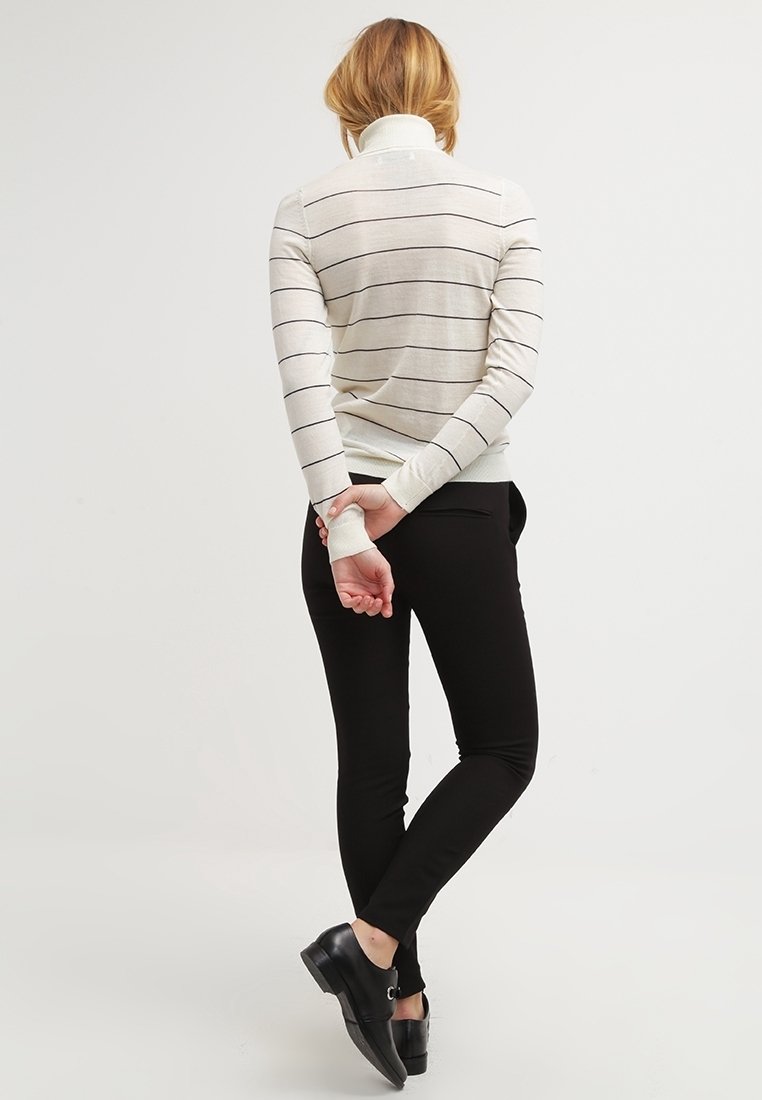 Krémový svetr s černými pruhy, rolák, žebrované manžety; kombinovaný s těsnými černými kalhotami a černými botami. Pohled zezadu.