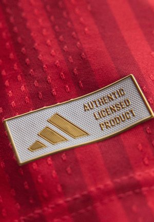 Bílá a zlatá etiketa na červené tkanině zobrazující tři diagonální pruhy a text "Autentický licencovaný produkt."
