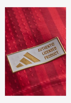 Bílá a zlatá etiketa na červené tkanině zobrazující tři diagonální pruhy a text "Autentický licencovaný produkt."