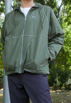 Veste coupe-vent vert olive avec des garnitures blanches, fermeture éclair intégrale, capuche ajustable et poches latérales, associée à un pantalon noir.