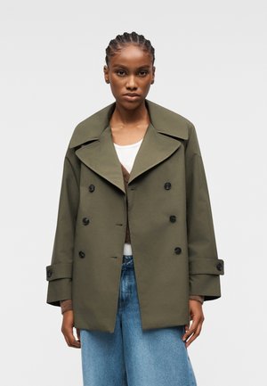 Kvinde med fletninger iført en oversized olivengrøn dobbeltradet frakke, hvid top, brun cardigan og lyseblå jeans.