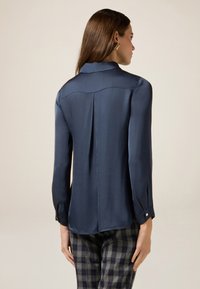 Blusa lunga in satin navy con colletto rotondo, dettaglio delle pieghe posteriori e polsini con bottoni, abbinata a pantaloni a quadri.