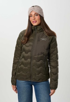 Une femme porte une veste matelassée vert olive à col montant, un bandeau en maille beige et un jean bleu, sur un fond uni.