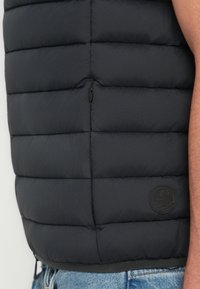 Sort polstret vest med kviltet design, der har en sidelomme med lynlås og et cirkulært logo-nål. Glat tekstur og letvægtsmateriale.