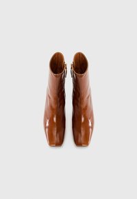 Botas de tobillo de cuero marrón con un acabado brillante, que presentan una punta cuadrada y detalles de cremallera dorada en el lateral. Vista superior.