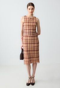 Frau trägt ein ärmelloses midi Kleid in Beige und Braun mit Karomuster, hält eine kleine dunkle gewebte Handtasche und trägt dunkle T-Strap High Heels.
