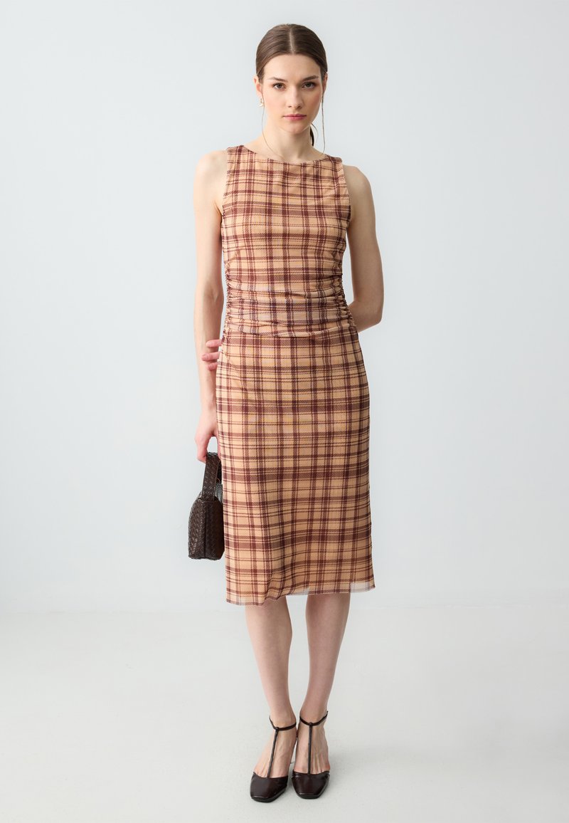 Frau trägt ein ärmelloses midi Kleid in Beige und Braun mit Karomuster, hält eine kleine dunkle gewebte Handtasche und trägt dunkle T-Strap High Heels.