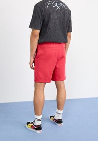 Červené bavlněné šortky s elastickým pasem, kombinované s tmavým oversized tričkem s bílým textem a černými teniskami s barevnými akcenty.
