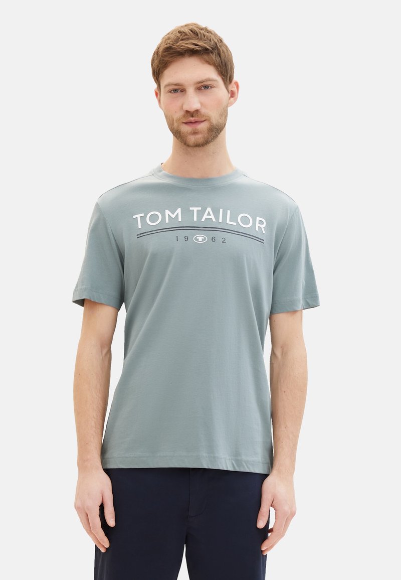 Camiseta de algodón gris con cuello redondo y mangas cortas, con "TOM TAILOR 1962" impreso en blanco contrastante a lo largo del pecho.