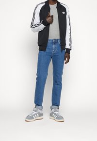 Černobílá bunda na zip s třemi pruhy, šedé tričko, modré džíny a šedé tenisky s bílými akcenty.