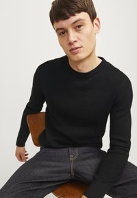 Svart ribbad tröja med långa ärmar och rund halsringning, tillsammans med mörka jeans som har synliga sömnadsdetaljer.