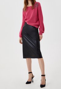 Žena v růžovém svetru s dlouhými rukávy a ozdobami, s černou koženou sukní po kolena, doplněná černými vysokými podpatky.