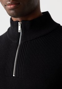 Black ribbed sweater with a high collar and silver zipper. Textured knit fabric creates visible vertical lines. Close-up view.
