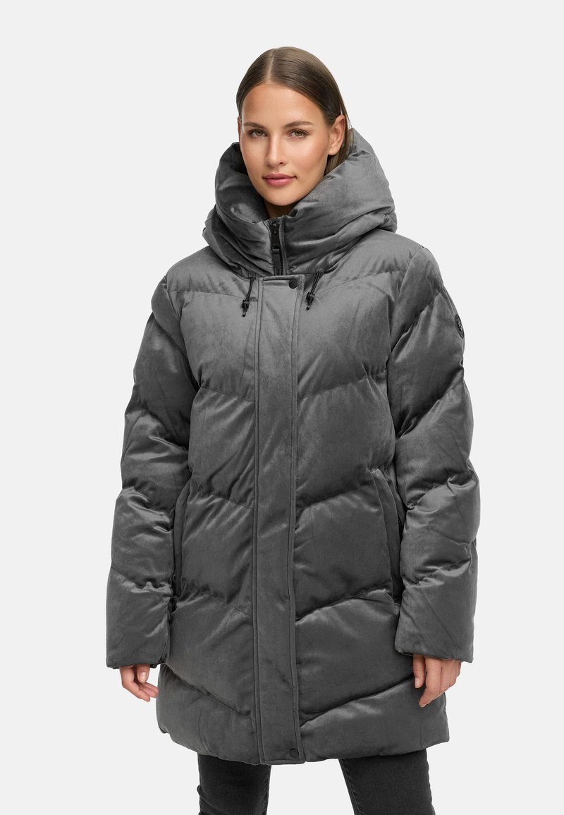 Sieviete ģērbusies pelēkā siltajā ziemas mētelī ar augstu, pildītu apkakli un rāvējslēdzēja aizdari, stāv pretī vienkāršai gaišai fona.