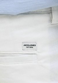 Bílý látkový kapesníček s našitou štítkem nesoucím nápis "JACK & JONES EST. 1990." Pozadí tvoří světle modrá látka.