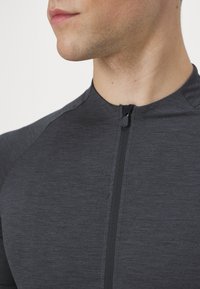 Haut de sport gris avec un col haut, fermeture éclair et tissu texturé, présentant des accents contrastants aux épaules. Gros plan sur la zone du col.